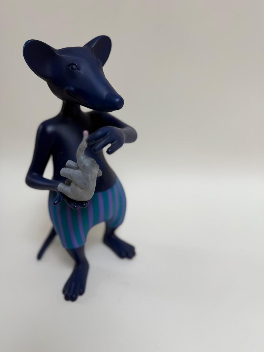 Souris stylisée debout, corps bleu et rayures vertes, tenant un objet dans ses pattes.